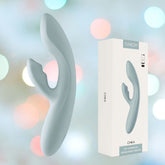 A stylish Svakom Chika Rabbit Vibrator in turquoise grey, designed for app control, is showcased alongside its packaging. It offers dual stimulation and is crafted from premium liquid silicone. The background features softly blurred colorful bokeh lights, adding to its charm.